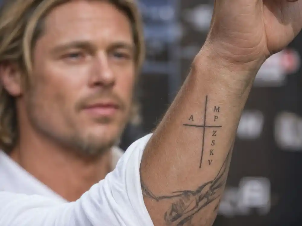 Brad Pitt tattoos and their meanings family cross — wrist cross tattoo with letter A and children initials M P Z S K V on forearm at premiere