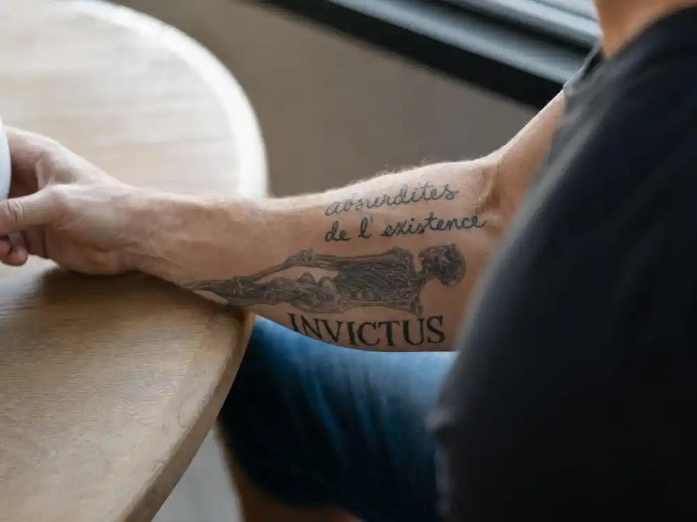 Brad Pitt tattoos and their meanings Ötzi Invictus — left forearm close-up with Iceman mummy silhouette Latin Invictus and French absurdites script