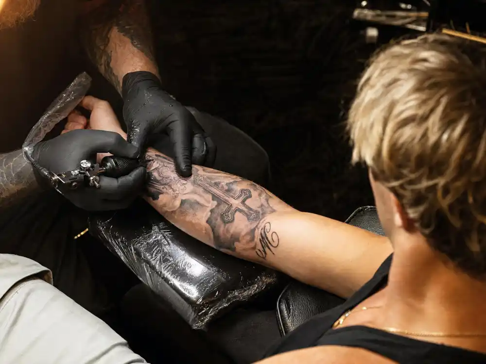 Brad Pitt tattoos and their meanings studio session — tattoo artist gloved hands applying ink machine to forearm cross design over-shoulder angle