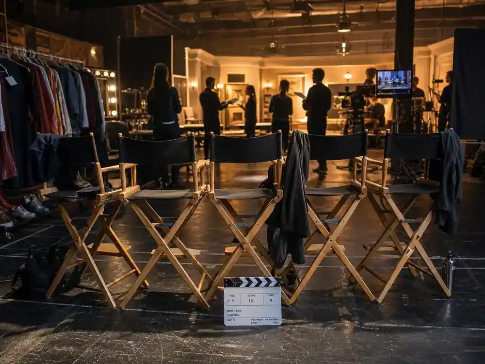 Breaking Bad cast net worth 2026 ranked ensemble — five director chairs in a row on TV drama soundstage with wardrobe racks and rehearsing actor silhouettes
