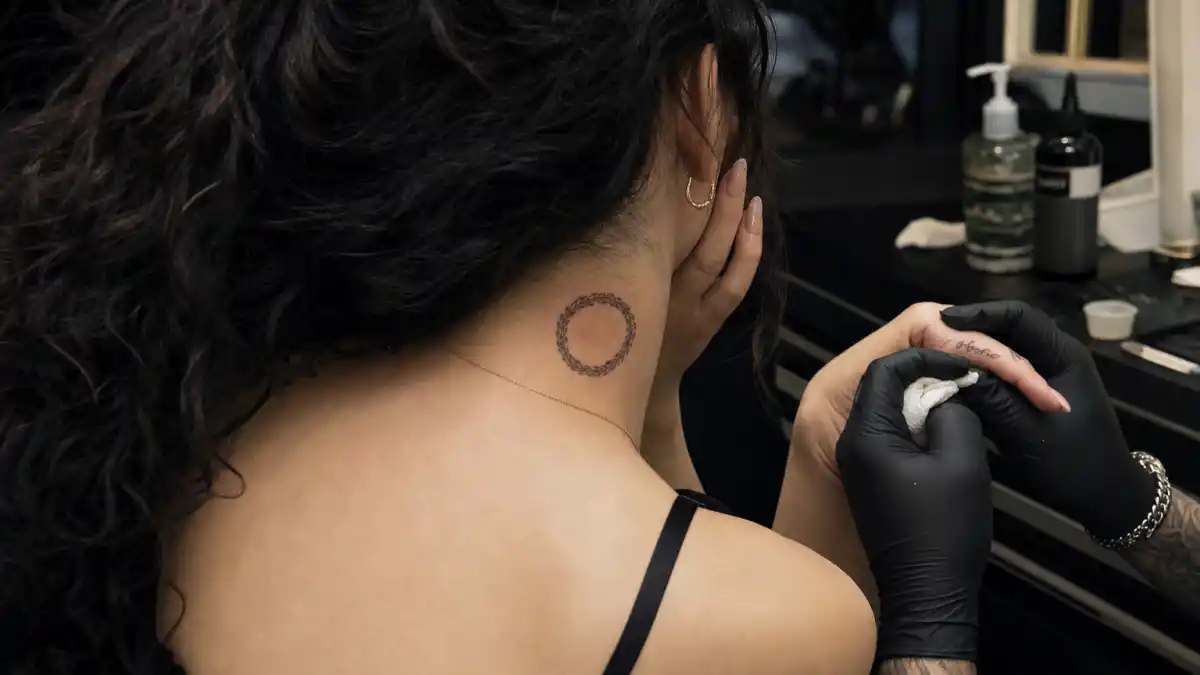 Camila Cabello tattoos and their meanings — petite woman with long dark wavy hair from behind at tattoo studio with pinky script and neck sweetgrass braid visible