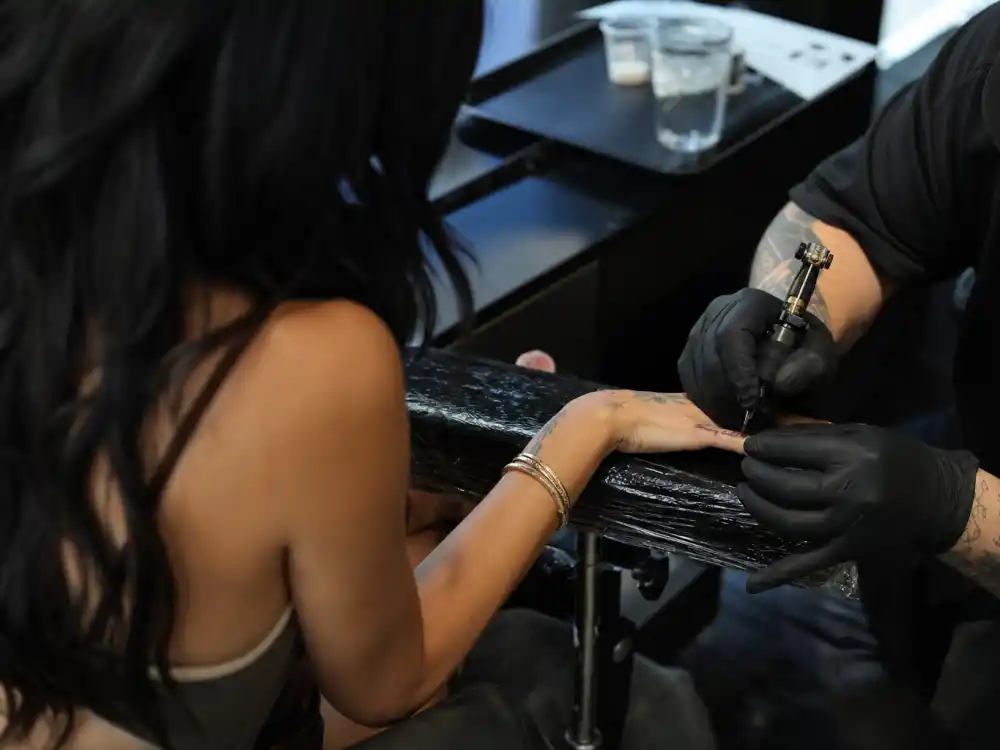 Camila Cabello tattoos and their meanings Kane Navasard — LA tattoo studio session with artist gloved hands applying fine-line pinky script