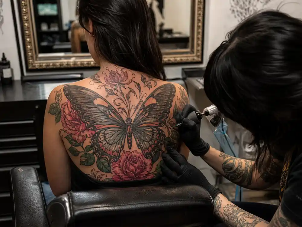 Cardi B tattoos and their meanings back flowers — tattoo artist working on massive butterfly and floral piece spanning upper back during Jamie Schene session