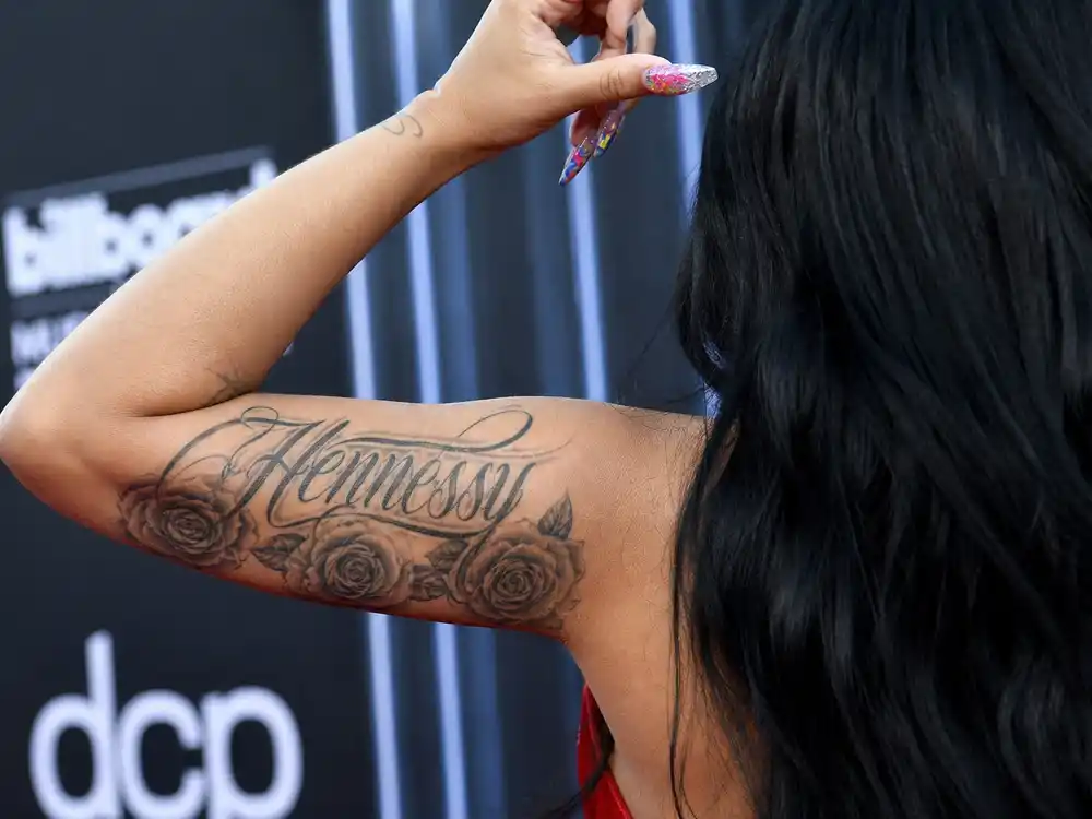 Cardi B tattoos and their meanings Hennessy — left bicep sister tribute script with three rose flowers backstage over-shoulder close-up