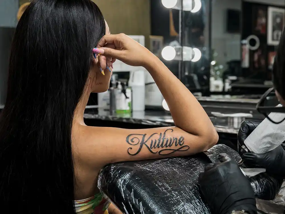 Cardi B tattoos and their meanings Kulture and Wave — inner bicep daughter name script and pale red son name jawline tattoo at mirror