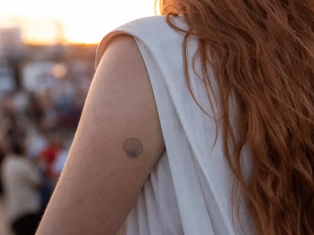 Chappell Roan tattoos and their meanings shell — small seashell tattoo above elbow on upper arm over-shoulder at outdoor golden hour
