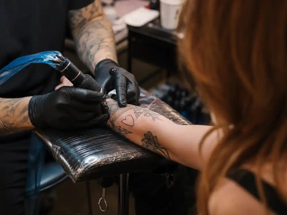 Chappell Roan tattoos and their meanings studio session — tattoo artist gloved hands applying floral ink on red-haired client forearm