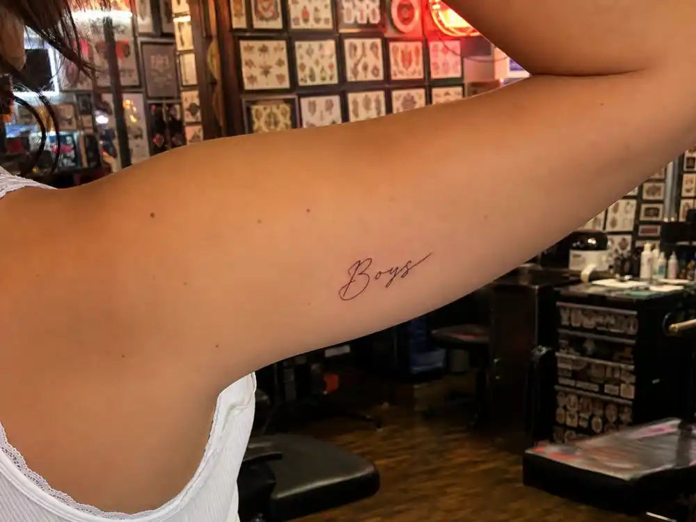 Charli XCX tattoos and their meanings — close-up of delicate cursive Boys script tattoo on upper arm from over-shoulder angle