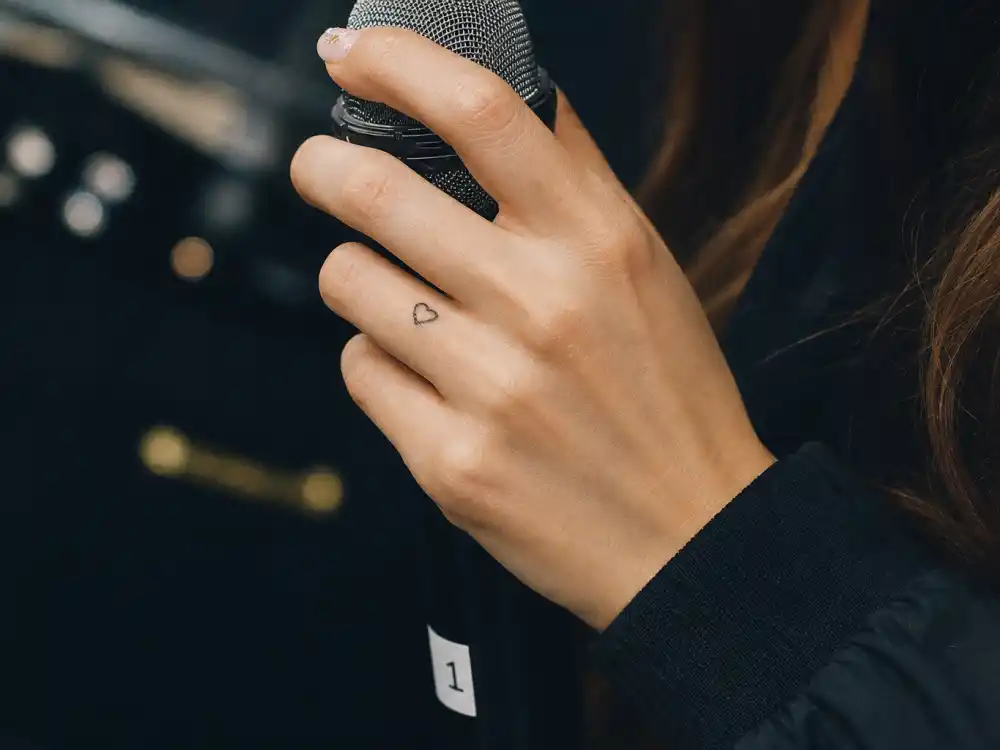 Charli XCX tattoos and their meanings — small minimalist heart tattoo on ring finger photographed backstage with microphone