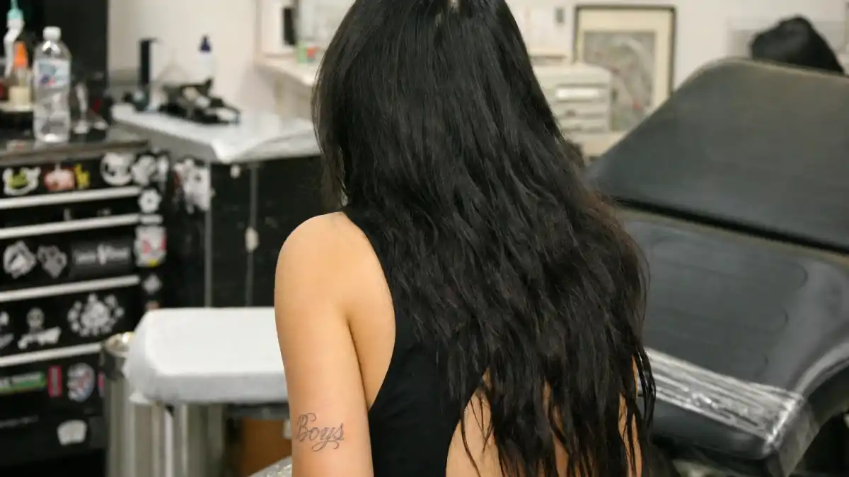 Charli XCX tattoos and their meanings — pop singer seen from behind in tattoo studio with delicate Boys cursive arm tattoo visible