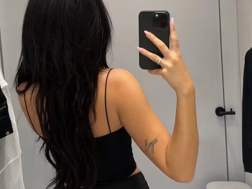 Charli XCX tattoos and their meanings — mirror angle showing Boys arm tattoo and heart finger ink in minimalist dressing room
