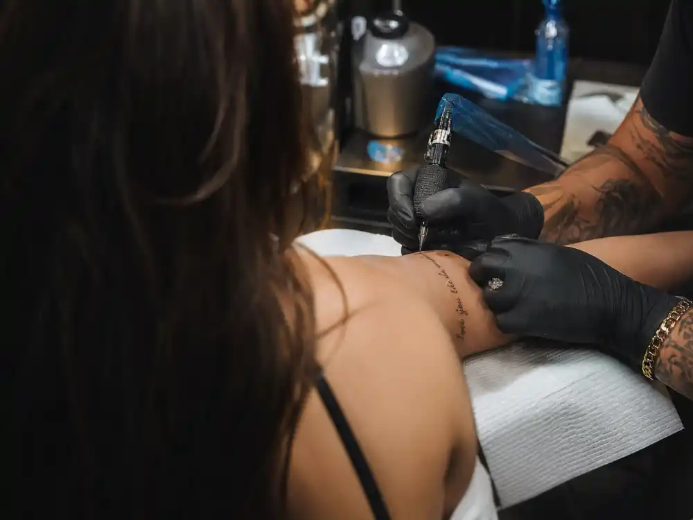 Charli XCX tattoos and their meanings — tattoo artist inking fine-line script during professional studio session from behind