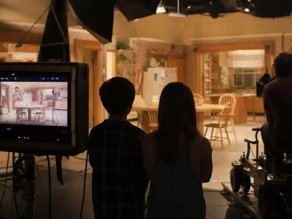 child actors from 90s sitcoms cast reunion guide child cast — young actor silhouettes from behind on 1990s suburban kitchen TV set with director monitor and camera dolly