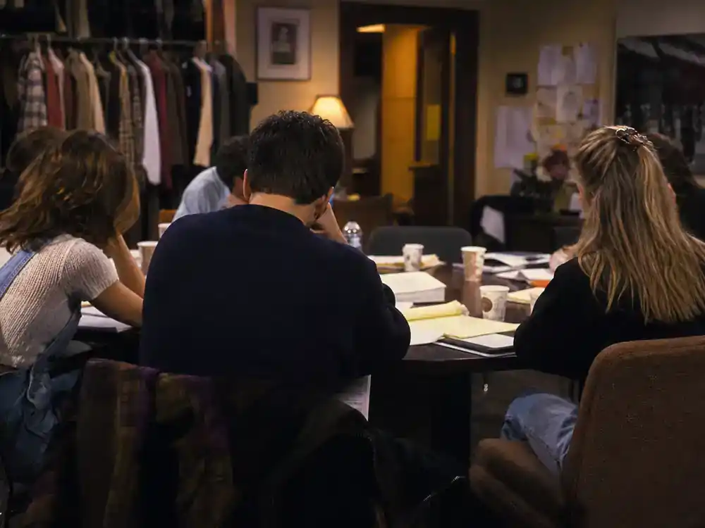 child actors from 90s sitcoms cast reunion guide sitcom sets — young TV comedy actor silhouettes from behind at table read with scripts and coffee cups in production office