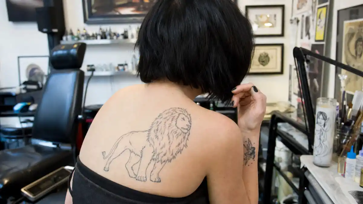 Christina Ricci tattoos and their meanings — actress seen from behind in tattoo studio with Aslan lion shoulder tattoo and fairy wrist ink visible
