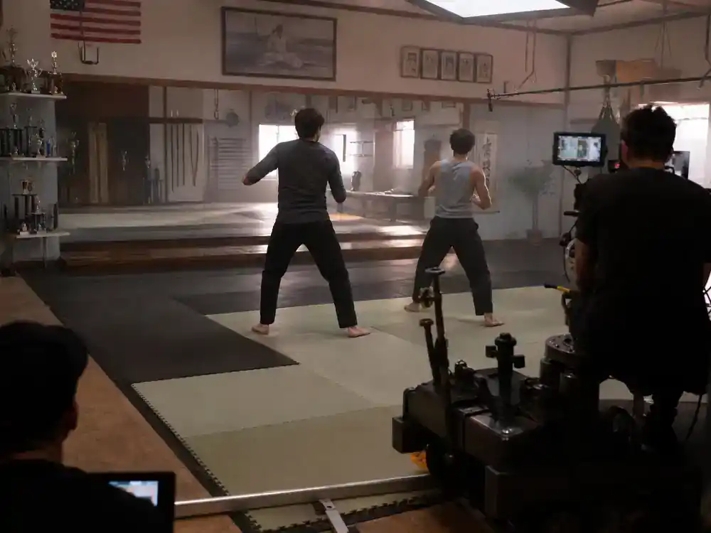 Cobra Kai cast and karate kid timeline guide Daniel LaRusso — two actor silhouettes from behind rehearsing blocking on suburban dojo film set with mirror wall