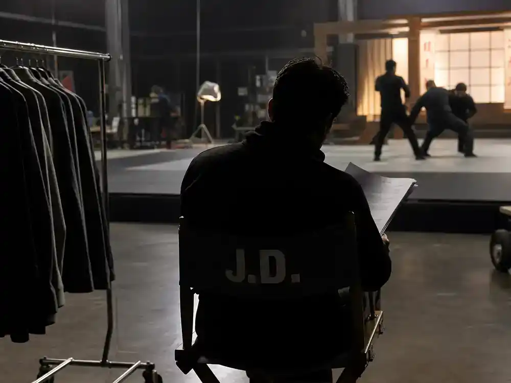 Cobra Kai cast and karate kid timeline guide Johnny Lawrence — lead actor silhouette from behind on director chair reviewing script at martial arts TV drama soundstage