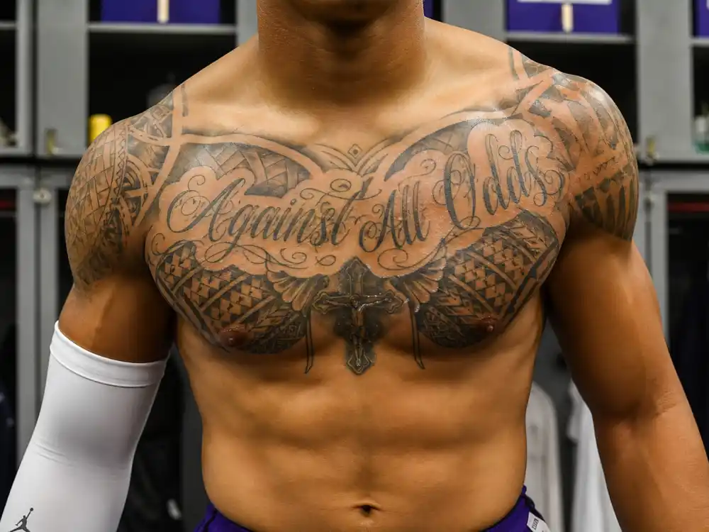 Colin Kaepernick tattoos and their meanings — Against All Odds chest script surrounded by Polynesian tribal patterns on both pecs