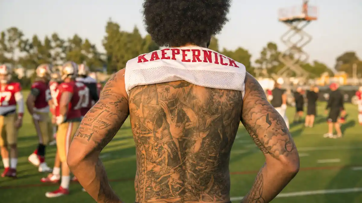 Colin Kaepernick tattoos and their meanings — quarterback seen from behind on practice field with angels-demons back mural and arm religious ink visible