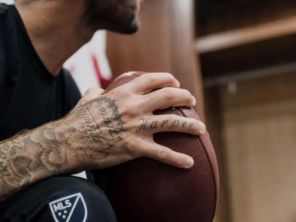 David Beckham tattoos and their meanings Harper hand — script finger tattoo on tattooed hand holding football in locker room close-up