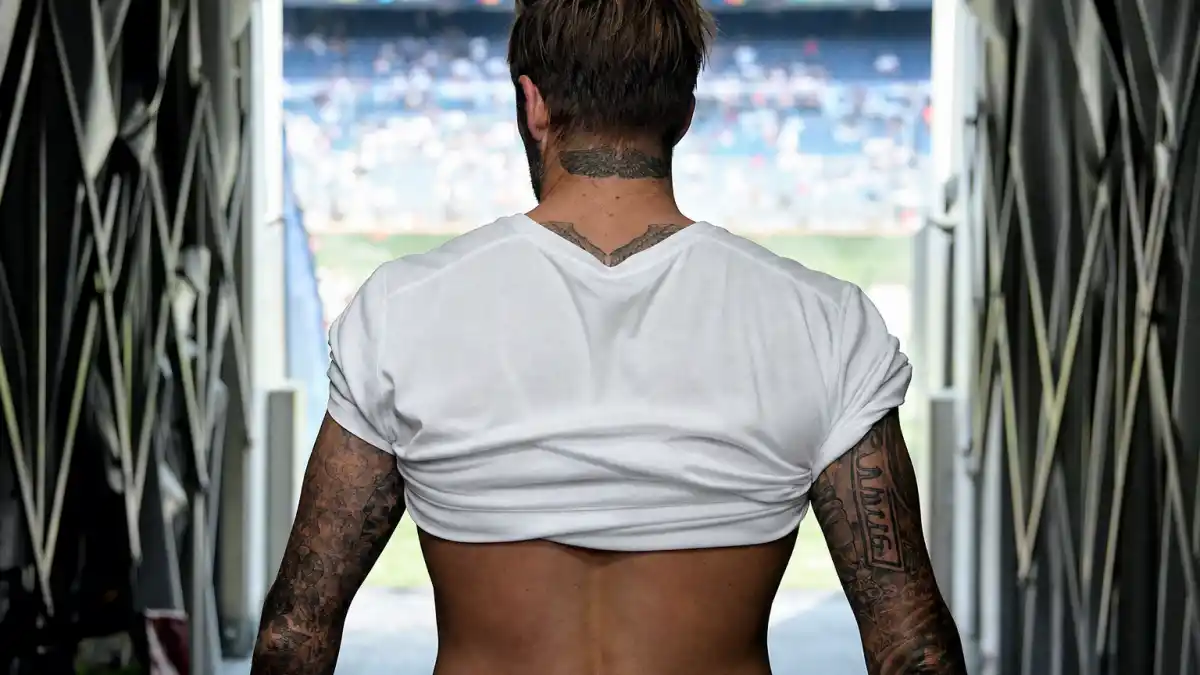 David Beckham tattoos and their meanings — athletic man with short dark hair from behind in stadium tunnel showing extensive tattoo sleeves on both forearms