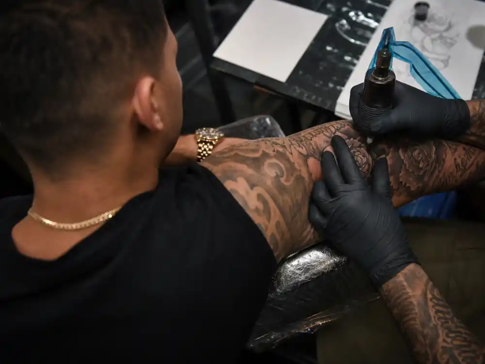 David Beckham tattoos and their meanings studio session — tattoo artist gloved hands applying ink machine to rose script forearm sleeve over-shoulder angle