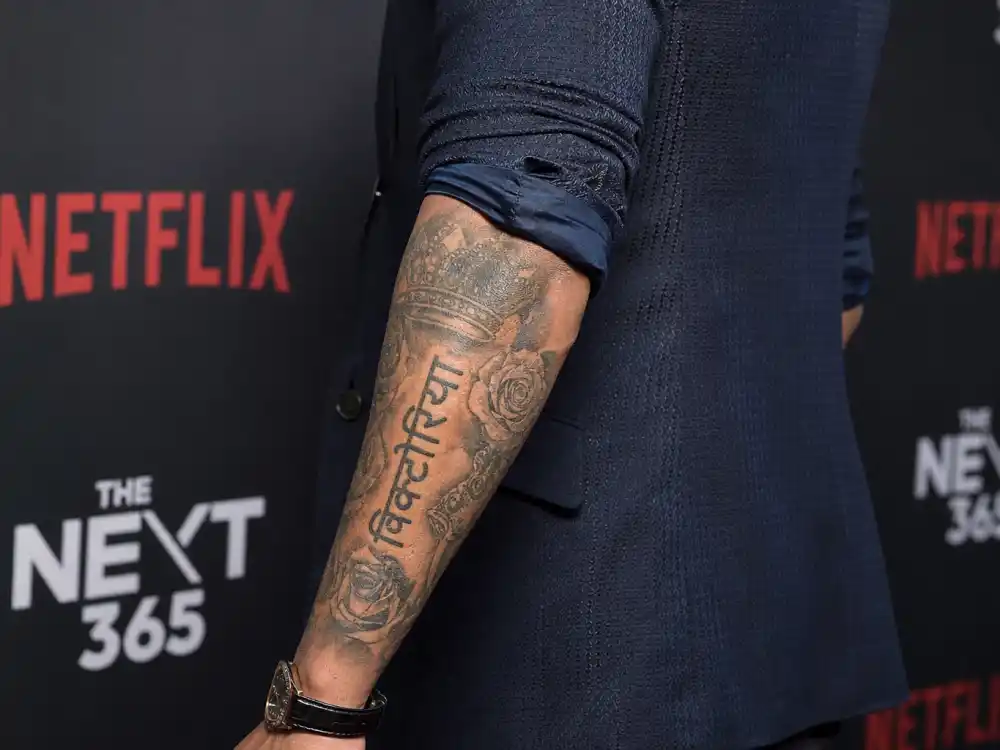 David Beckham tattoos and their meanings Victoria script — Hindi Devanagari Victoria tattoo with roses on forearm sleeve at red carpet over-shoulder angle