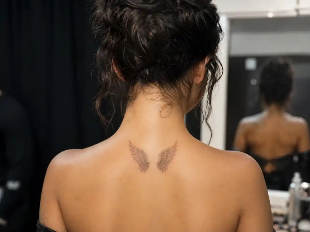 Demi Lovato tattoos and their meanings — angel wings tattoo on back of neck representing spiritual rebirth in dressing room mirror