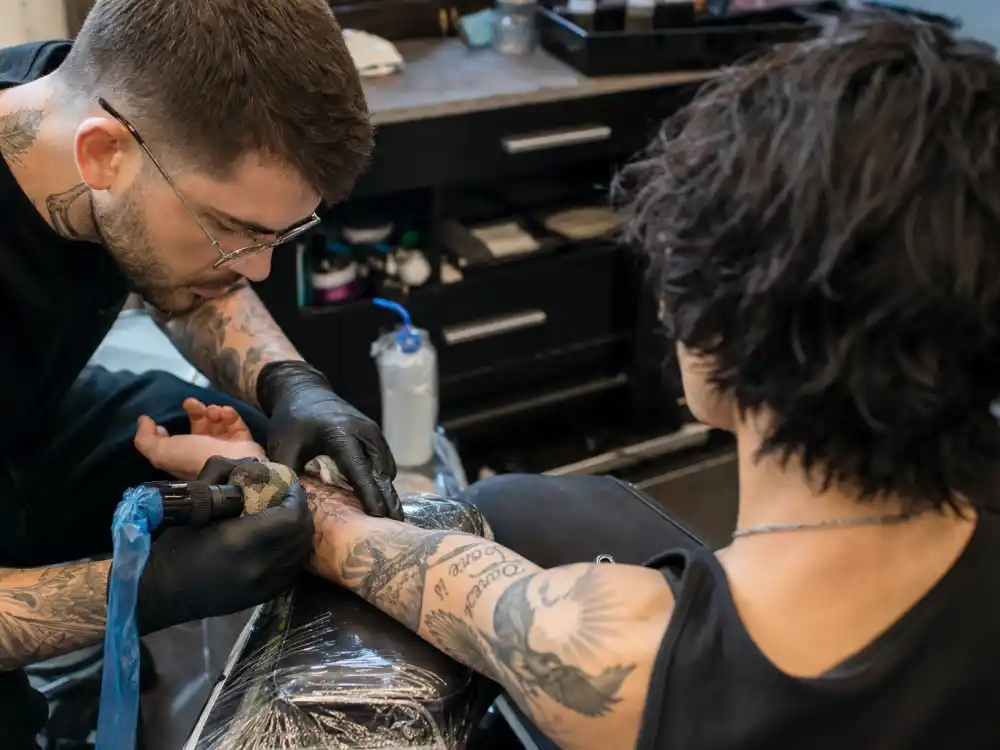Demi Lovato tattoos and their meanings — tattoo artist inking forearm during professional studio session with existing bird and script tattoos visible