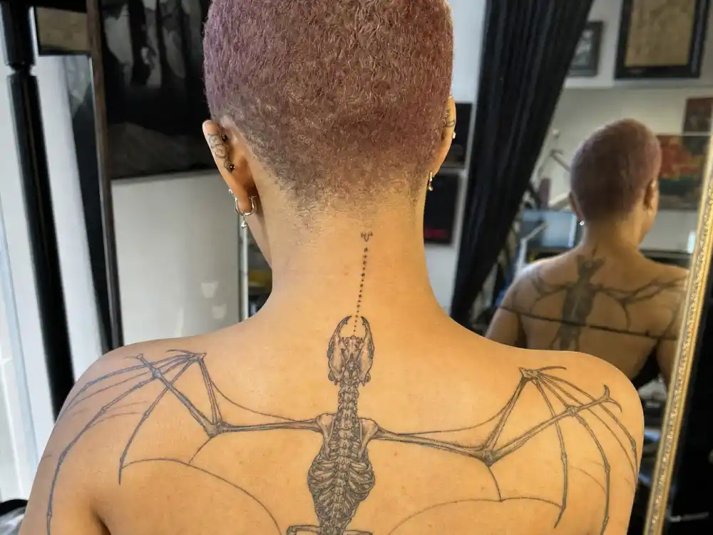 Doja Cat tattoos and their meanings — bat skeleton fine-line tattoo spanning upper back and shoulders photographed from behind in studio mirror