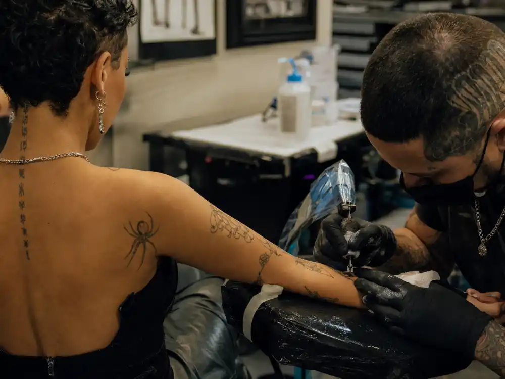 Doja Cat tattoos and their meanings — tattoo artist inking forearm during professional studio session with existing spider and fine-line tattoos visible