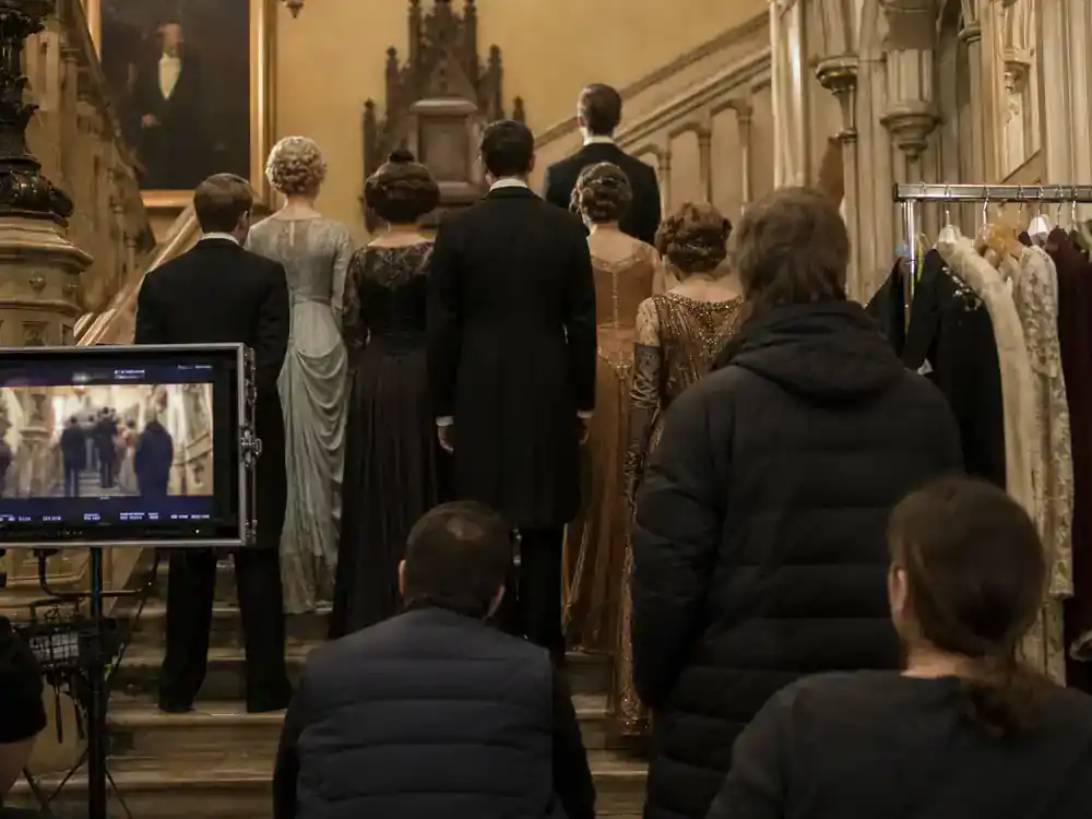 Downton Abbey cast and movie timeline guide Crawley family — upstairs aristocracy TV drama cast silhouettes from behind on ornate manor staircase set with Edwardian costumes and director monitor