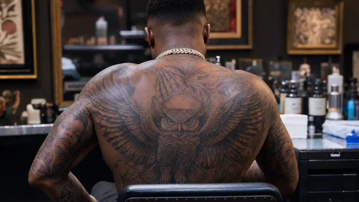 Drake tattoos and their meanings — hip-hop artist seen from behind in tattoo studio with massive owl back piece and full arm sleeves visible