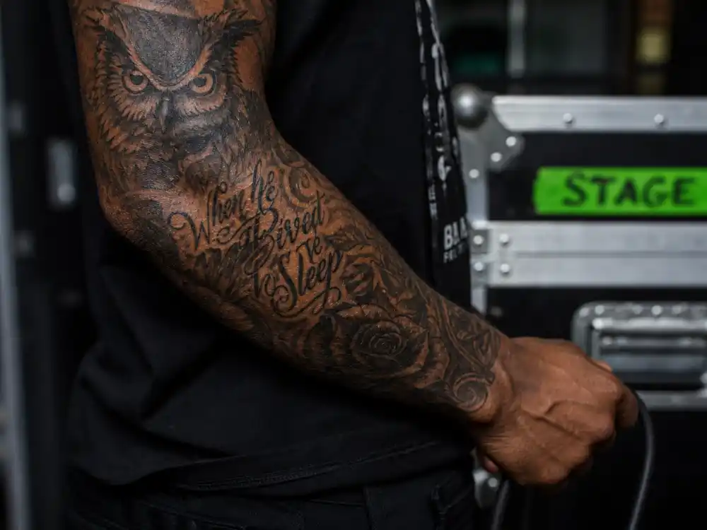 Drake tattoos and their meanings — close-up of heavily tattooed forearm sleeve with owl and script motifs on musician arm