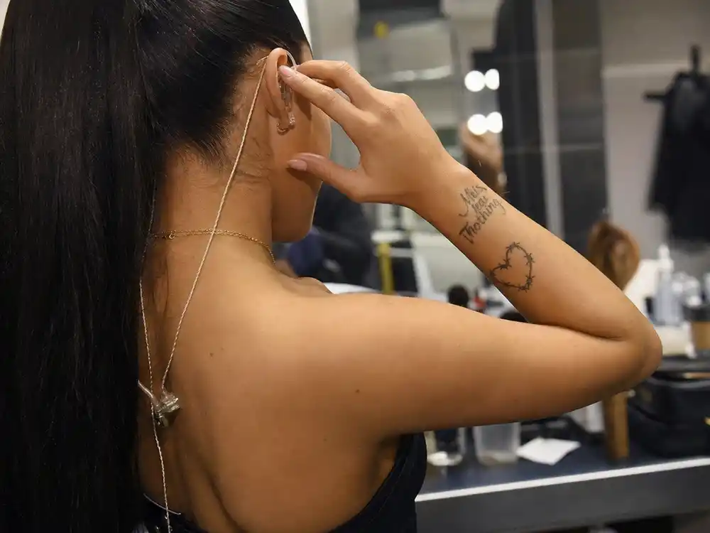 Dua Lipa tattoos and their meanings — This Means Nothing script and barbed wire heart forearm tattoos in backstage over-shoulder close-up