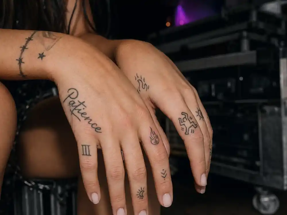 Dua Lipa tattoos and their meanings — patience hand script, flame index finger, and Keith Haring dancing figure thumb tattoos in fine black ink