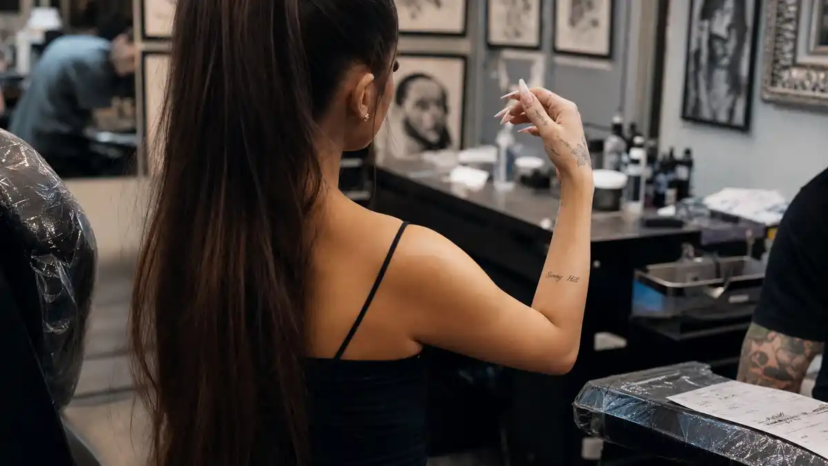 Dua Lipa tattoos and their meanings — pop star seen from behind in tattoo studio with Sunny Hill forearm script and delicate hand micro tattoos visible