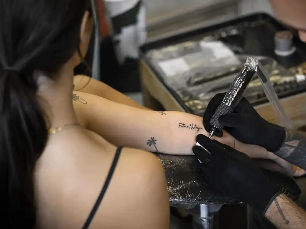 Dua Lipa tattoos and their meanings — tattoo artist inking forearm during professional studio session with Future Nostalgia script visible