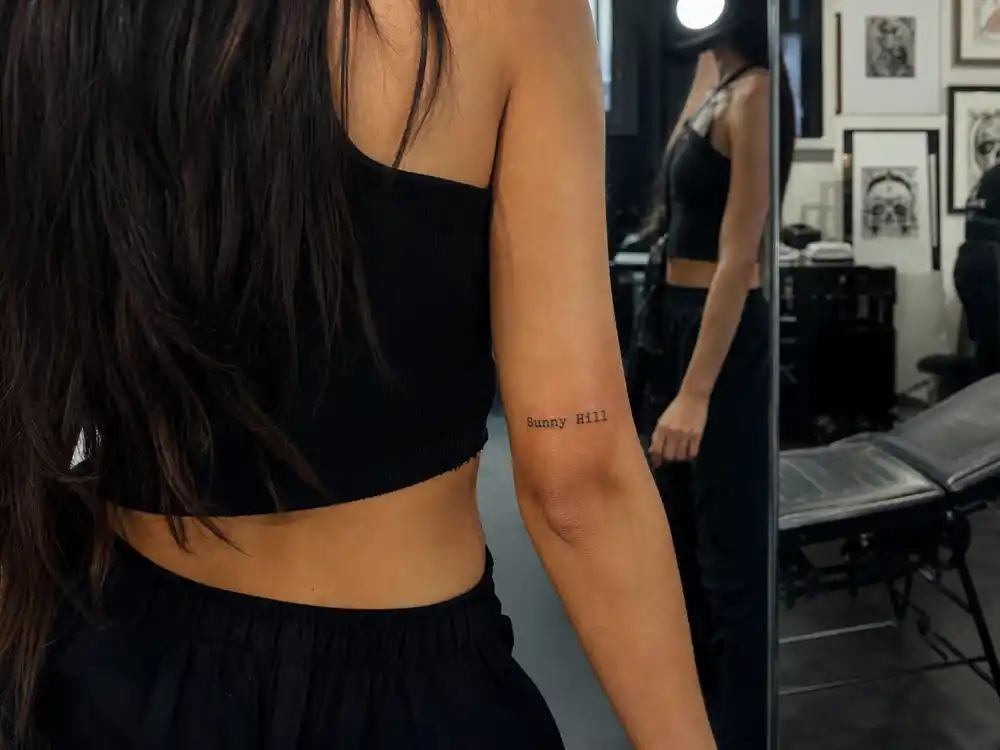 Dua Lipa tattoos and their meanings — Sunny Hill script tattoo on inner forearm photographed from behind in tattoo studio mirror
