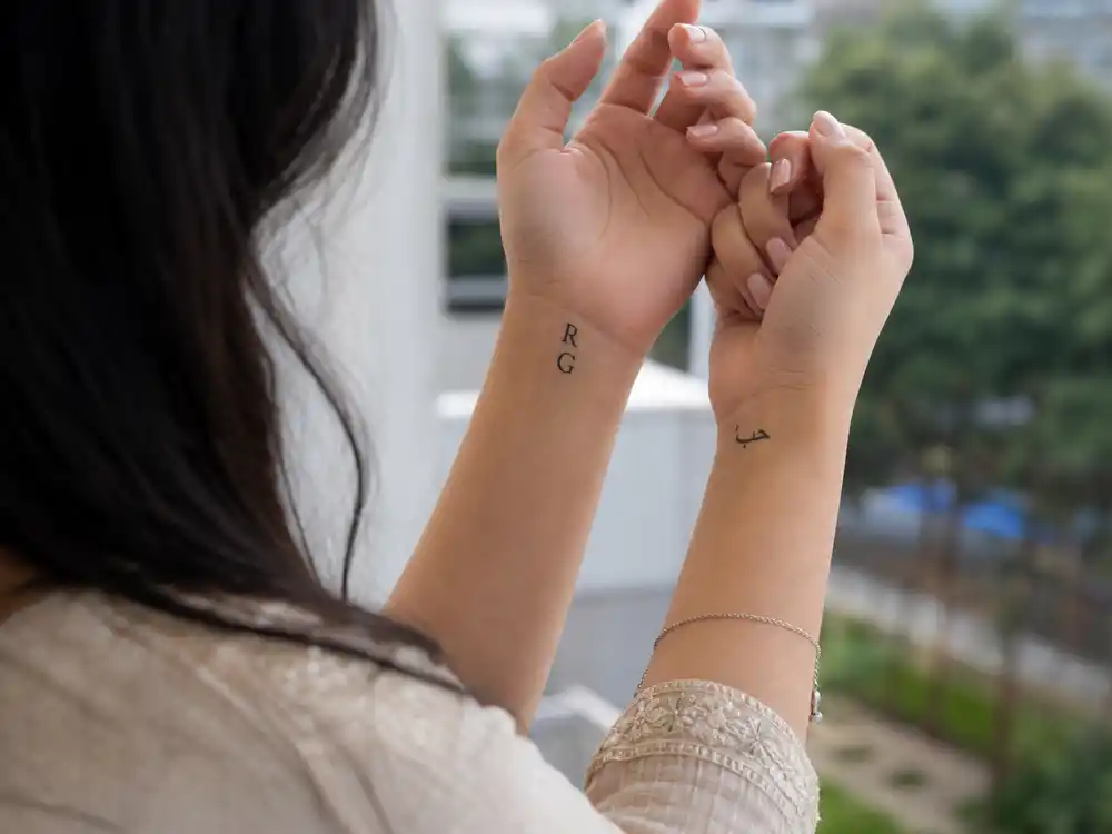 Dua Lipa tattoos and their meanings — R and G sibling initial tattoos and Arabic love script on wrists photographed over-shoulder