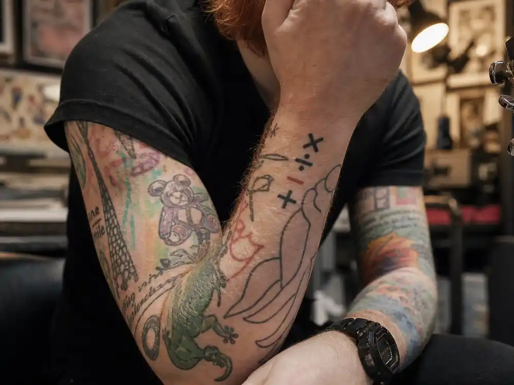 Ed Sheeran tattoos and their meanings arm ink — close-up of plus multiply divide subtract wrist symbols and colorful sleeve tattoos