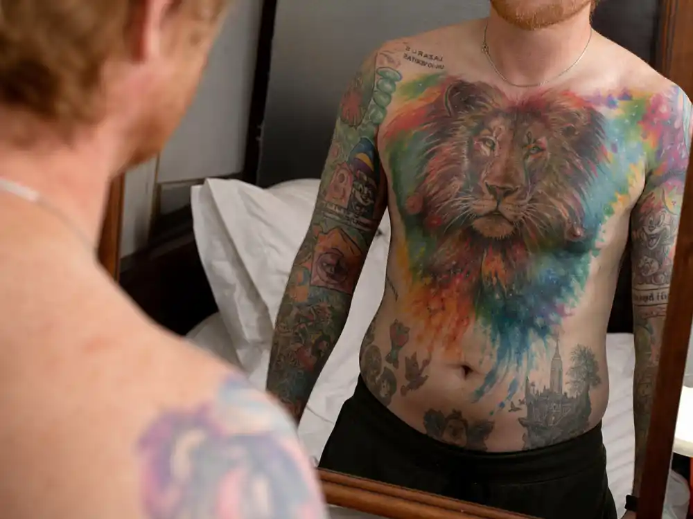 Ed Sheeran tattoos and their meanings chest piece — rainbow lion mural and colorful torso ink over-shoulder mirror angle