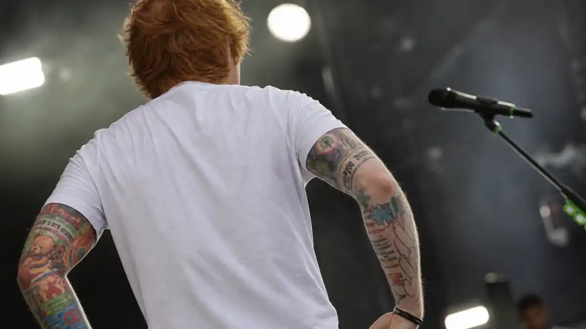 Ed Sheeran tattoos and their meanings — ginger-haired singer from behind showing colorful arm sleeve ink on concert stage