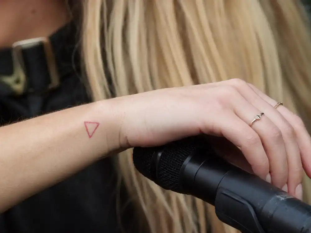 Ellie Goulding tattoos and their meanings triangle — red ink triangle outline tattoo on right wrist close-up holding microphone backstage
