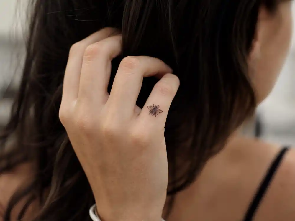 Emilia Clarke tattoos and their meanings bumblebee — tiny black bumblebee tattoo on left pinky finger honoring Me Before You role