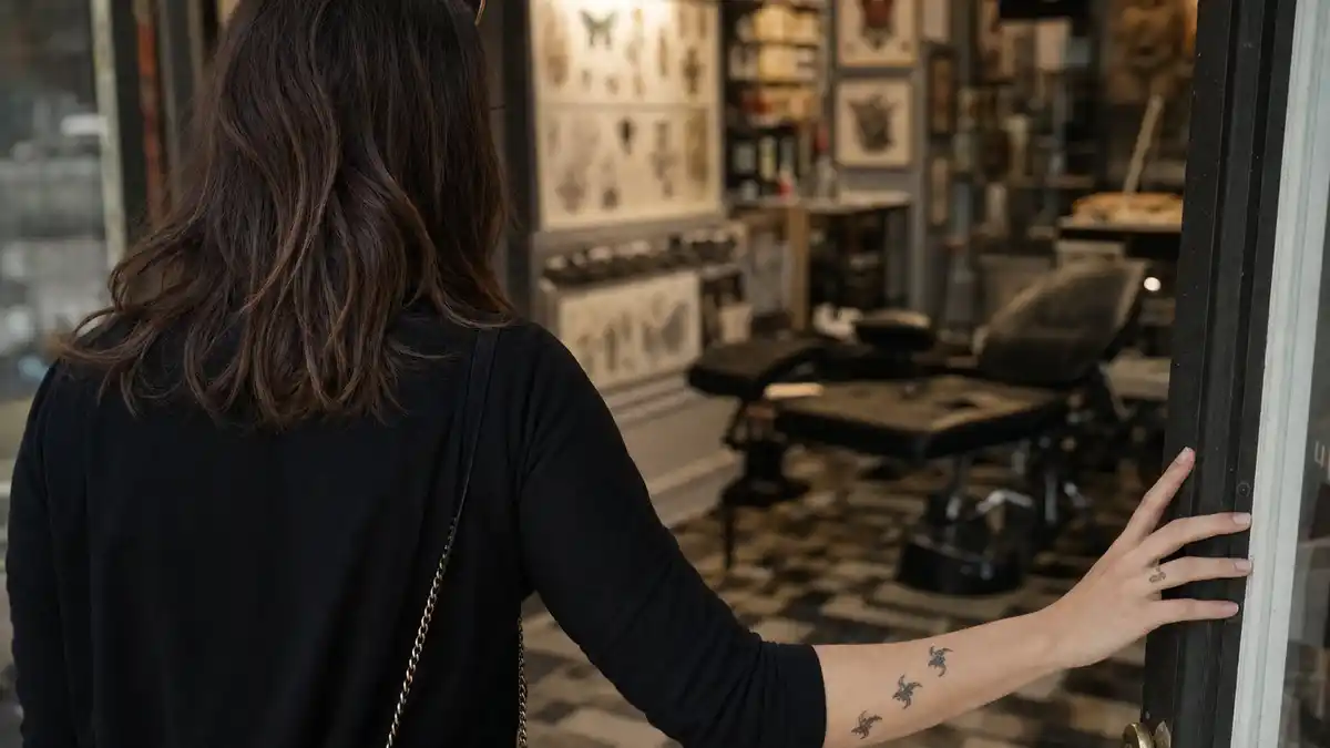 Emilia Clarke tattoos and their meanings — brunette actress from behind in tattoo studio showing delicate wrist dragon and pinky bumblebee tattoos