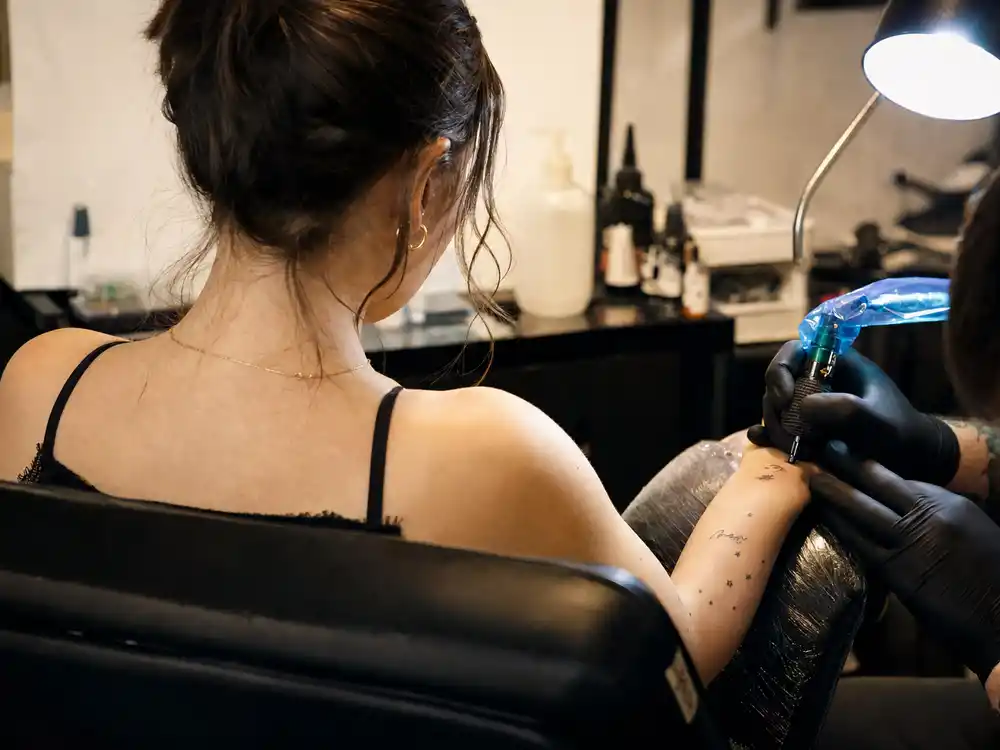 Emilia Clarke tattoos and their meanings studio session — petite brunette actress from behind in tattoo chair with wrist and finger ink visible
