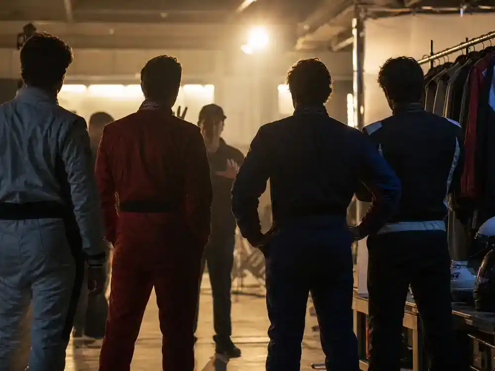 F1 movie Brad Pitt cast and racing facts ensemble cast — performer silhouettes in racing coveralls on soundstage pit garage set from behind