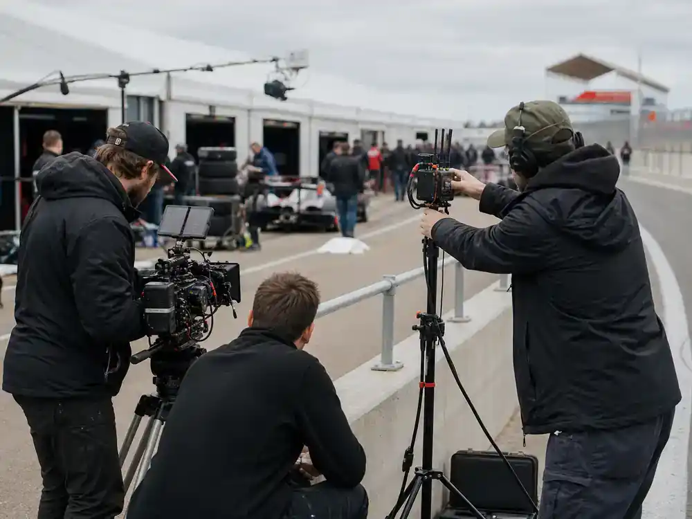 F1 movie Brad Pitt cast and racing facts filming — grand prix circuit paddock crew setting trackside cameras during real race weekend movie shoot