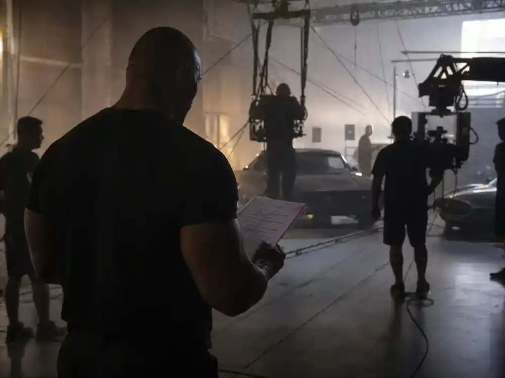 Fast and Furious movies cast order guide Dwayne Johnson — actor silhouette from behind reviewing scripts on warehouse stunt set with wire rig